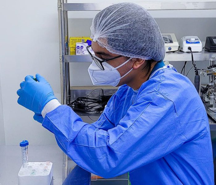 Hombre en un laboratorio, con mascarilla, anteojos, bata, guantes y gorra de laboratorio, observando una muestra.