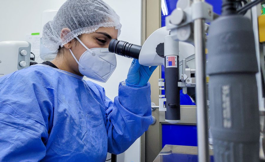 Mujer en un laboratorio, con mascarilla, bata, guantes y gorra de laboratorio, observando por el lente de un microscopio.