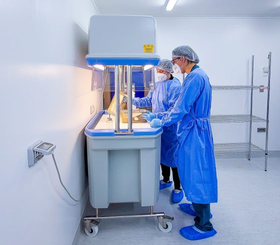 Dos personas con mascarilla, bata, guantes y gorra de laboratorio, observan una muestra de experimento en un laboratorio.